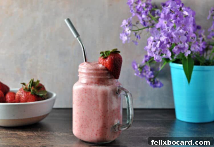 Keto strawberry smoothie with a straw, garnished with a fresh strawberries. Purple flowers and a bowl of fresh berries behind the smoothie.