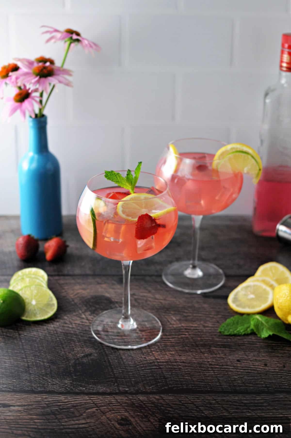 Blush Gin Sparkler 4 2 wine glasses of pink gin and lemonade cocktails surrounded by lemon and lime slices and a blue vase with flowers behind