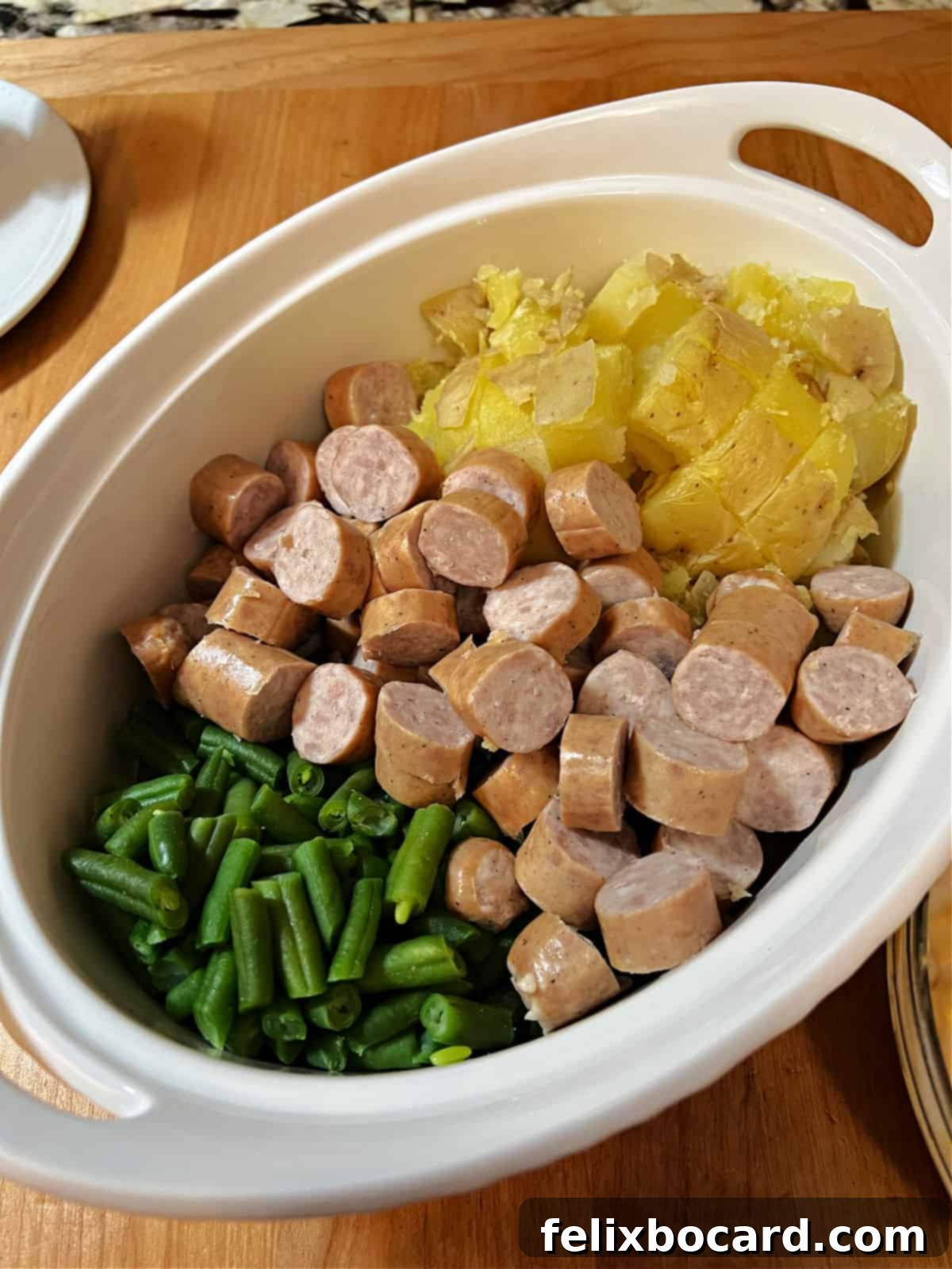 Casserole ingredients, including sliced bratwurst, cubed potatoes, and green beans, are neatly arranged in a baking dish, ready for the sauce and oven. This visually represents the initial assembly phase of the cheesy bratwurst stew.