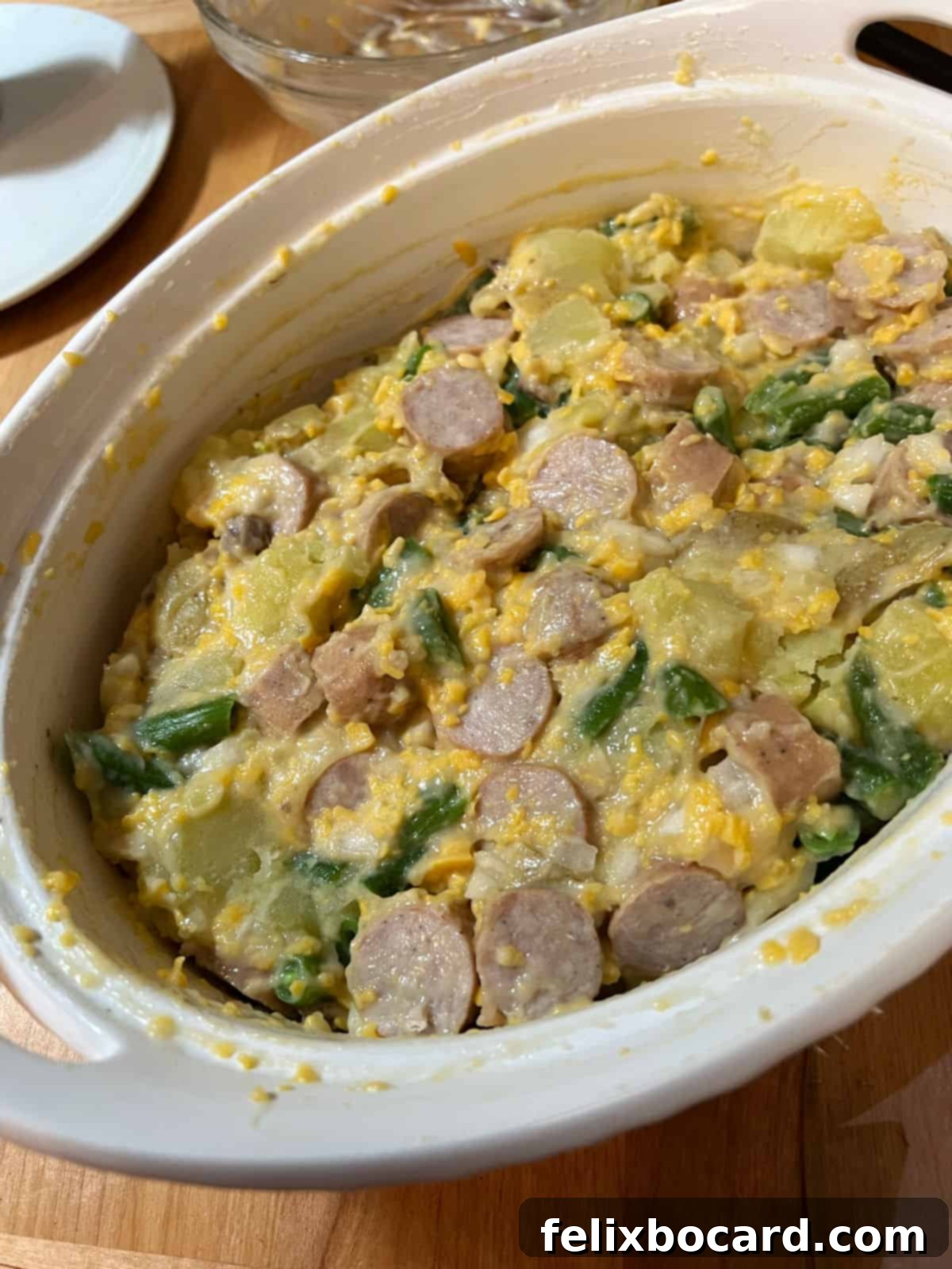 The assembled cheesy bratwurst casserole, with all ingredients mixed and ready for baking, sits in a baking dish. The rich, creamy sauce coats the bratwurst pieces, potatoes, and green beans, promising a delicious outcome.