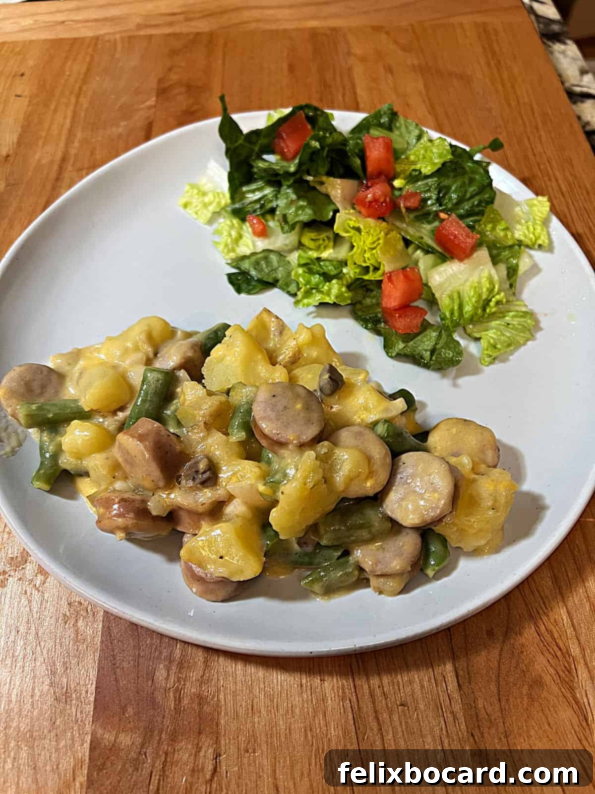 A beautifully plated serving of cheesy bratwurst casserole, accompanied by a vibrant green salad on the side. The casserole is golden and bubbly, highlighting its rich, comforting appeal.