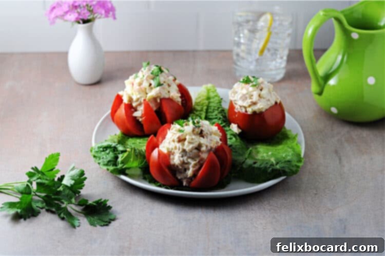 A vibrant plate featuring three tuna stuffed tomatoes, accompanied by a glass of water, a green pitcher, and a small vase of flowers in the background.