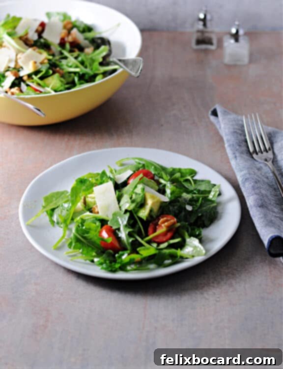 A bowl of freshly made Arugula Avocado Salad with a serving plate in front of it, displaying a portion of the salad.