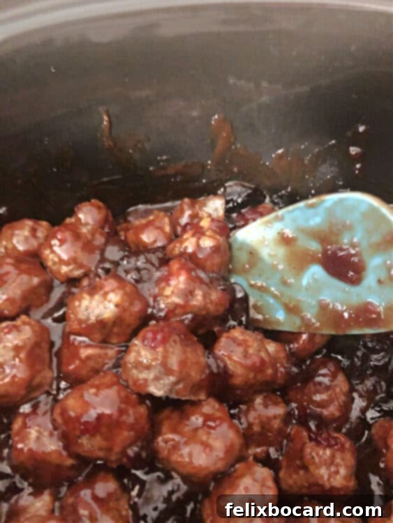 A spoon gently stirring meatballs in a crockpot, ensuring they are thoroughly coated in the bubbling grape jelly and BBQ sauce mixture.