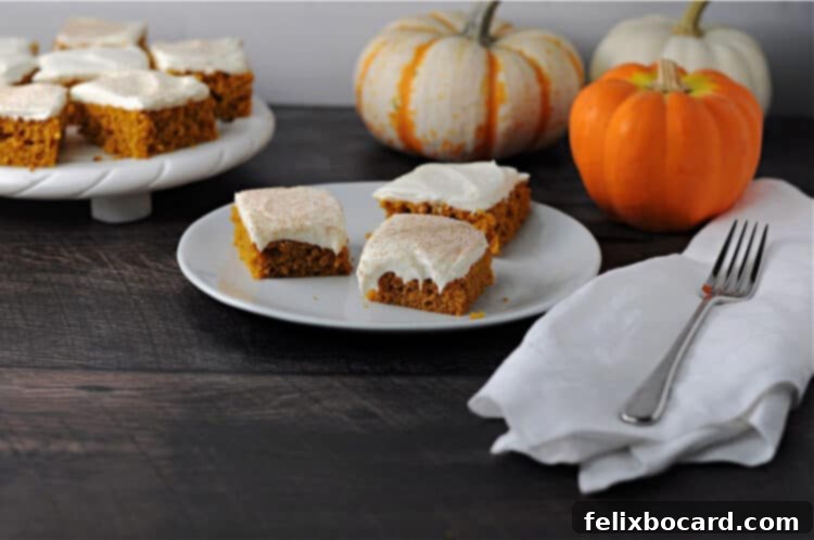 Three perfectly frosted pumpkin bars arranged on a white plate, with a pan of more bars visible in the background, highlighting the golden-orange hue and creamy white topping.