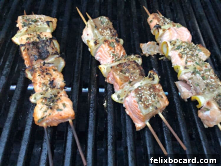 Several salmon kabobs arranged neatly on the hot grates of an outdoor grill, with visible char marks and steam rising, indicating they are actively cooking.