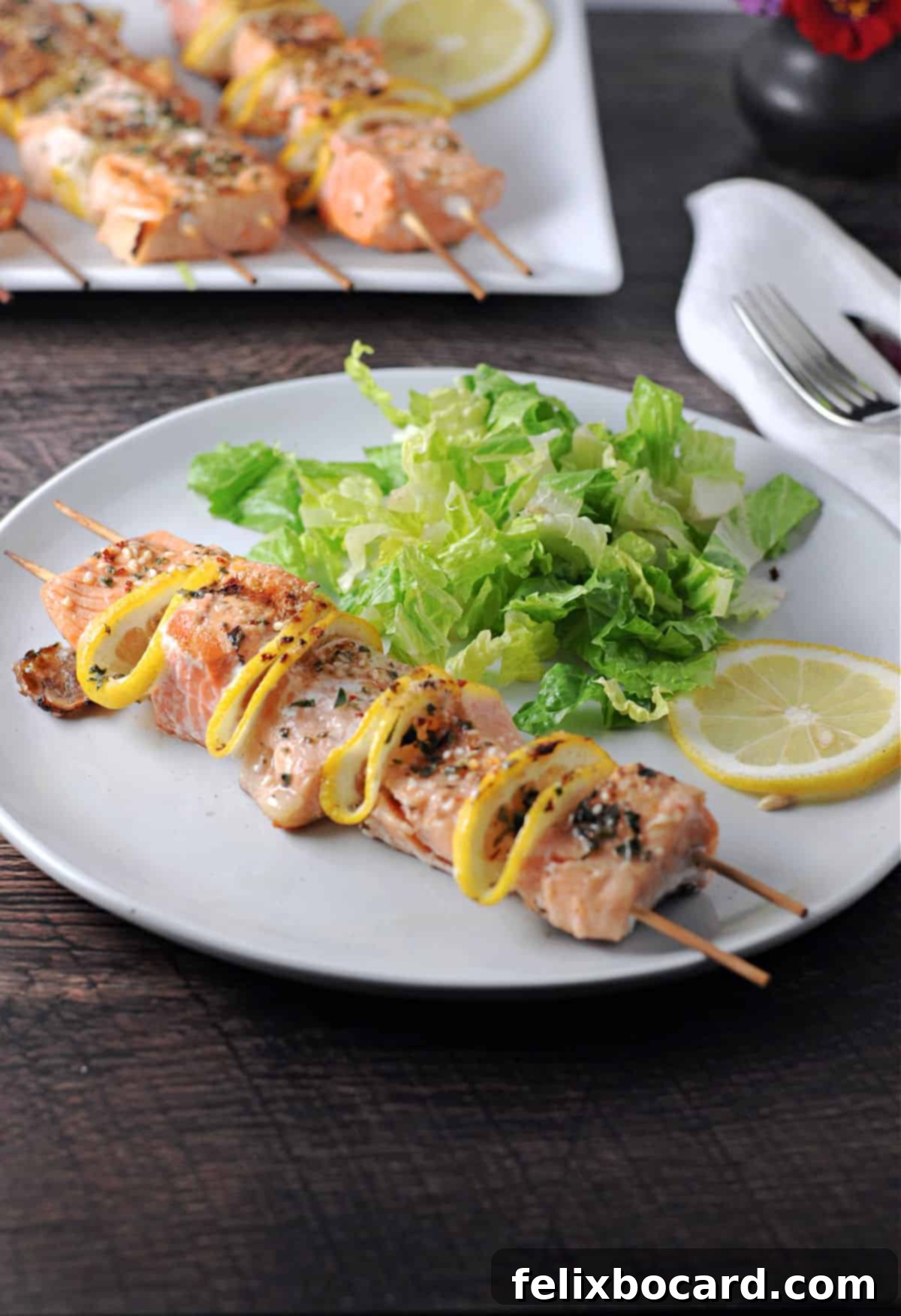 A vibrant close-up of a cooked salmon kabob resting on a white plate, accompanied by a fresh green salad. Additional kabobs are artfully blurred in the background, creating a mouth-watering scene.