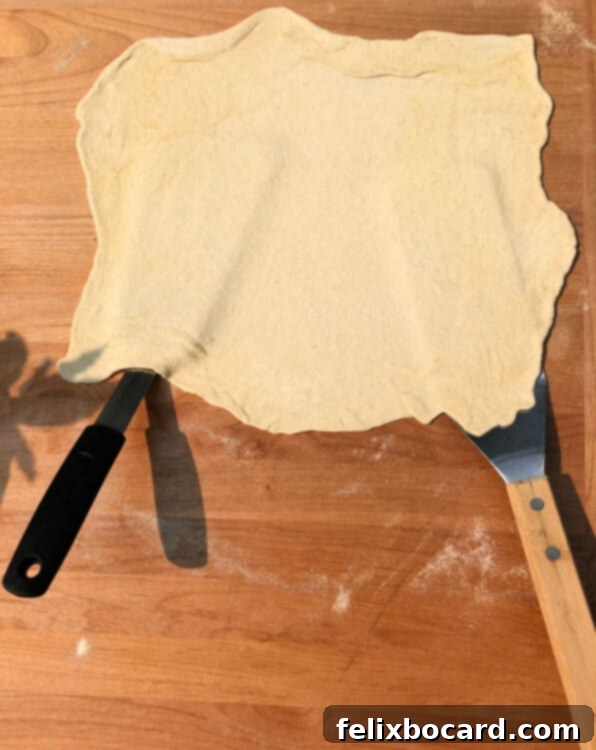 Two large spatulas picking up the delicate, rolled-out pizza dough from a cutting board, preparing to transfer it to a hot grill.