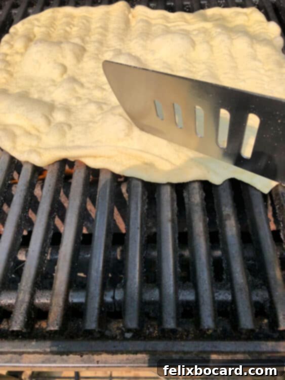 Spatula gently smashing down large bubbles in the grilling pizza crust to ensure even cooking and a flat surface.