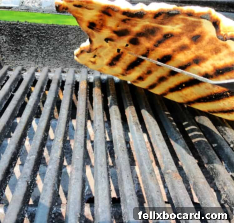 Grilled pizza crust lifted slightly to reveal the perfectly cooked underside with appealing char and grill marks, indicating it's ready to flip.
