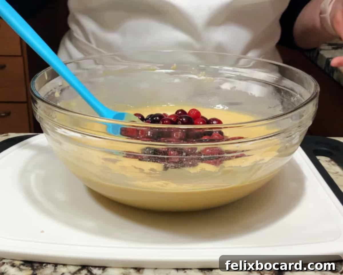 A bowl filled with a creamy batter, being mixed with a spatula, showing the rich texture before cranberries are added.