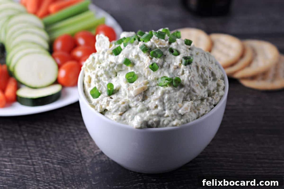 Bold Artichoke Jalapeno Dip 2 A bowl of creamy jalapeno artichoke dip, garnished, with a platter of crackers and fresh vegetables like cucumber slices and carrot sticks arranged behind it, ready for dipping.