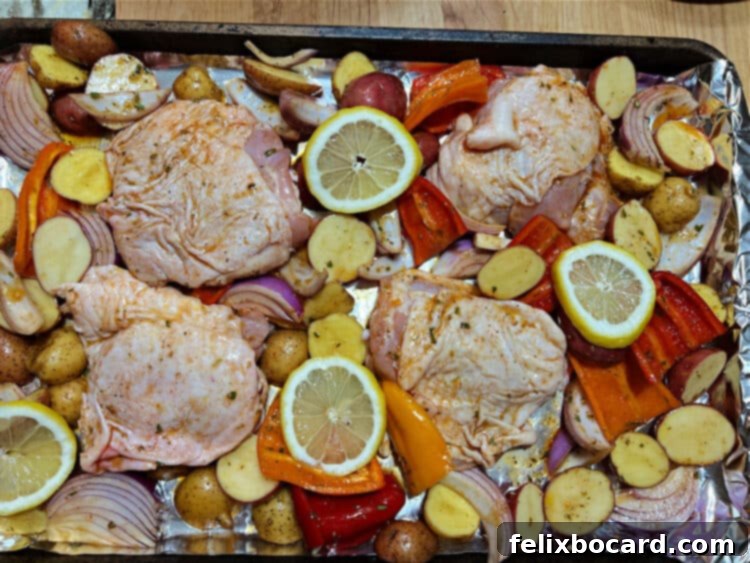 A sheet pan filled with seasoned Mediterranean chicken thighs, red onions, bell peppers, and baby potatoes, ready to be baked. Lemon slices are scattered among the vegetables, and the chicken skin is exposed for browning.
