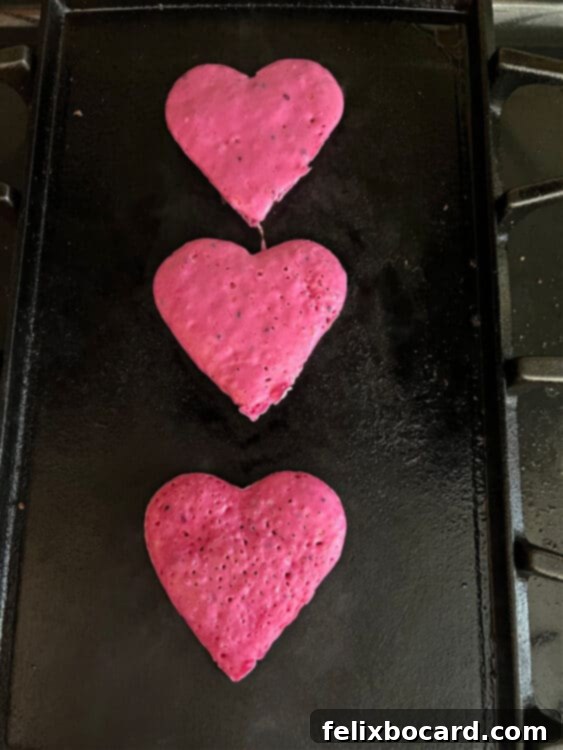 Three perfectly golden and vibrant pink heart-shaped dragon fruit pancakes cooking on a griddle, steam gently rising