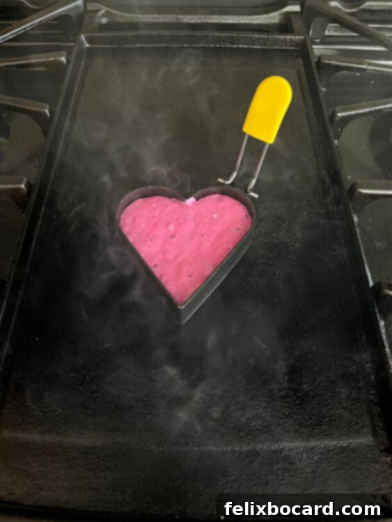 A heart-shaped mold resting on a hot griddle, filled with bright pink dragon fruit pancake batter