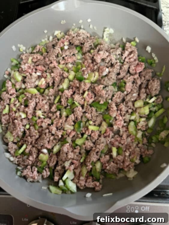 Cooked ground beef, now browned and crumbled, is mixed with softened chopped green pepper, celery, and onion in a skillet, indicating the initial cooking stage is complete.