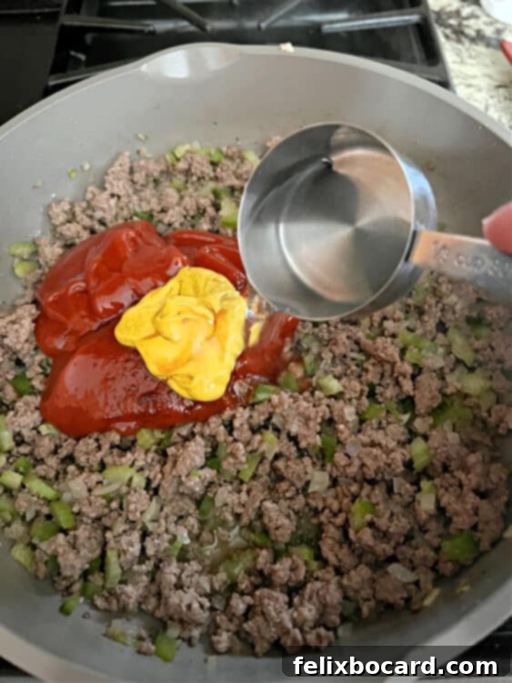 A hand is adding ketchup and other sauce ingredients directly into a skillet containing cooked ground beef and vegetables, beginning the sauce creation process.