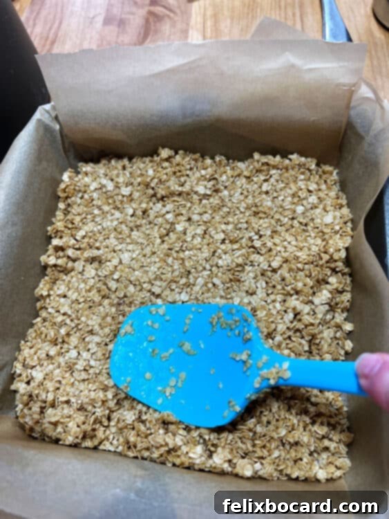The cooked oat mixture being pressed into a parchment-lined square pan with a spatula.
