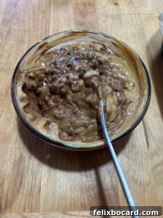 Partially melted chocolate chips being stirred into peanut butter in a glass bowl after microwaving.