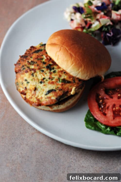 Top-down view of a grilled chicken feta spinach burger on a brioche bun, served on a white plate with fresh tomato slices, crisp lettuce, and a vibrant green salad on the side, ready for a healthy meal.