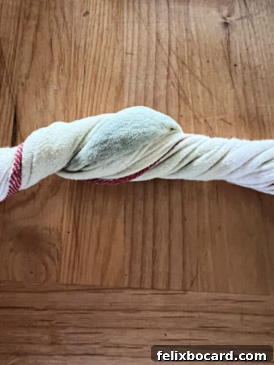 Close-up shot of a hand using a thin cotton towel to firmly squeeze excess moisture from thawed spinach over a kitchen sink. The spinach is tightly bundled in the towel, and drops of water are visible, highlighting the importance of this step for dry ingredients.