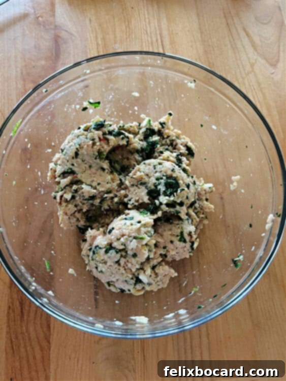 A close-up of a large mixing bowl filled with a fresh, uncooked chicken burger mixture, containing ground chicken, green spinach, and white crumbled feta cheese, thoroughly combined and ready to be formed into patties.