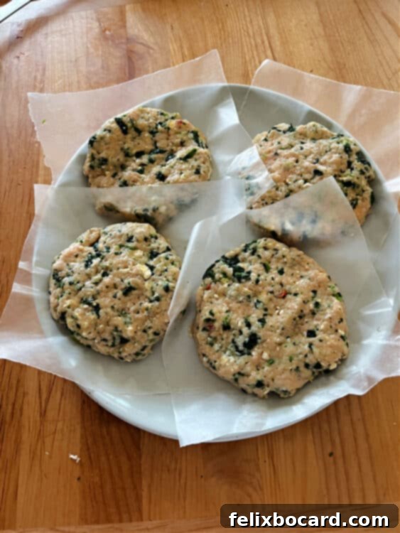 Four perfectly formed, uncooked chicken burger patties, made with ground chicken, feta, and spinach, neatly arranged on a plate lined with wax paper, ready for chilling before cooking.