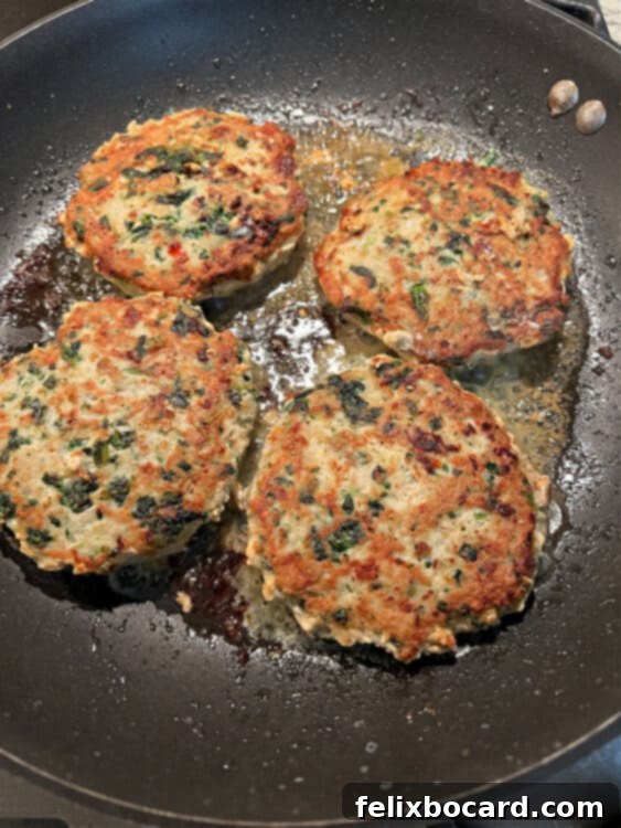 Three delicious chicken feta spinach burgers sizzling and cooking in a hot frying pan, with golden-brown crusts developing on their surfaces, indicating they are nearly ready to be served.