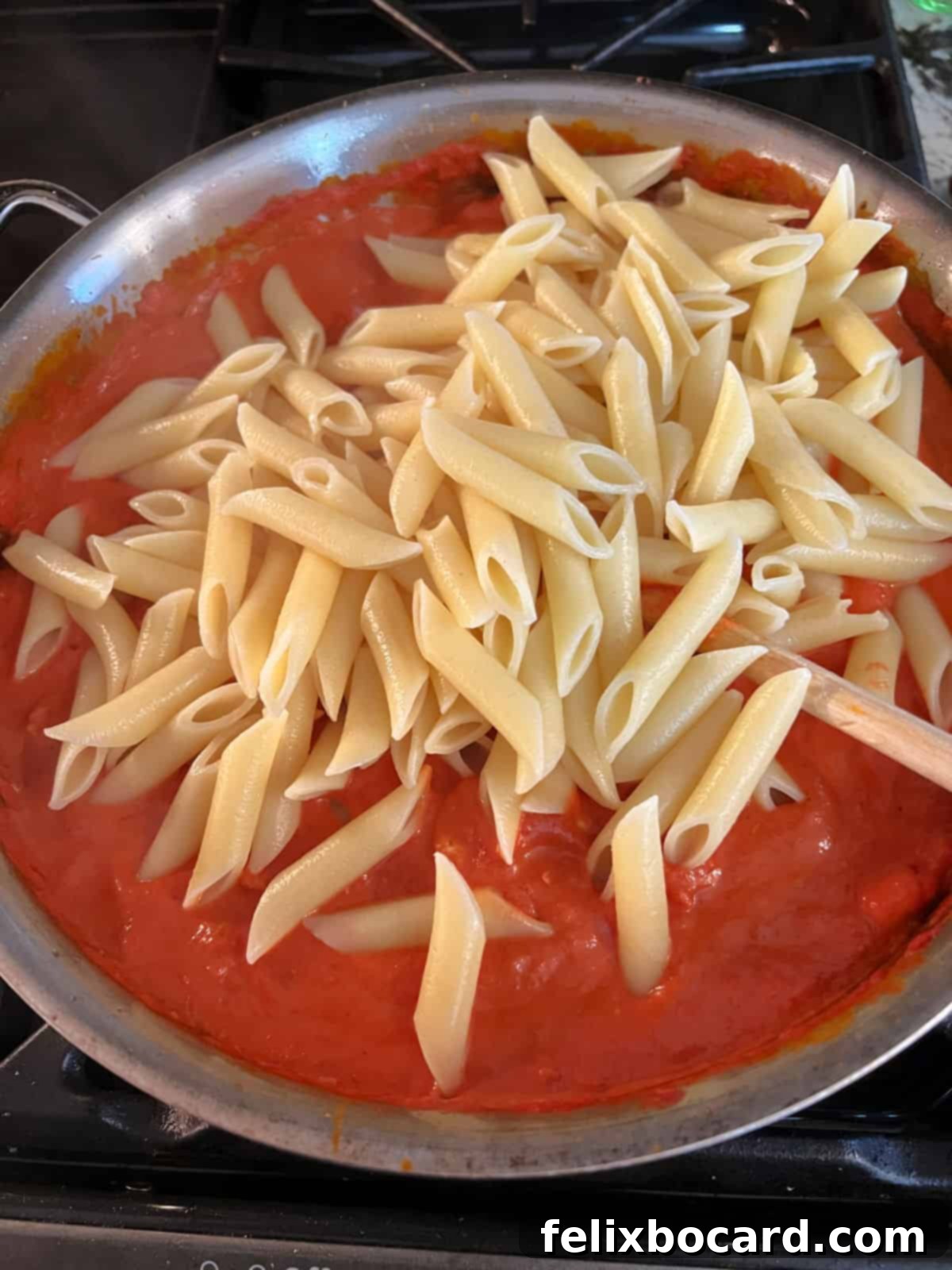 Cooked penne pasta being added directly from the pot into a large skillet filled with creamy vodka sauce, ready to be tossed.