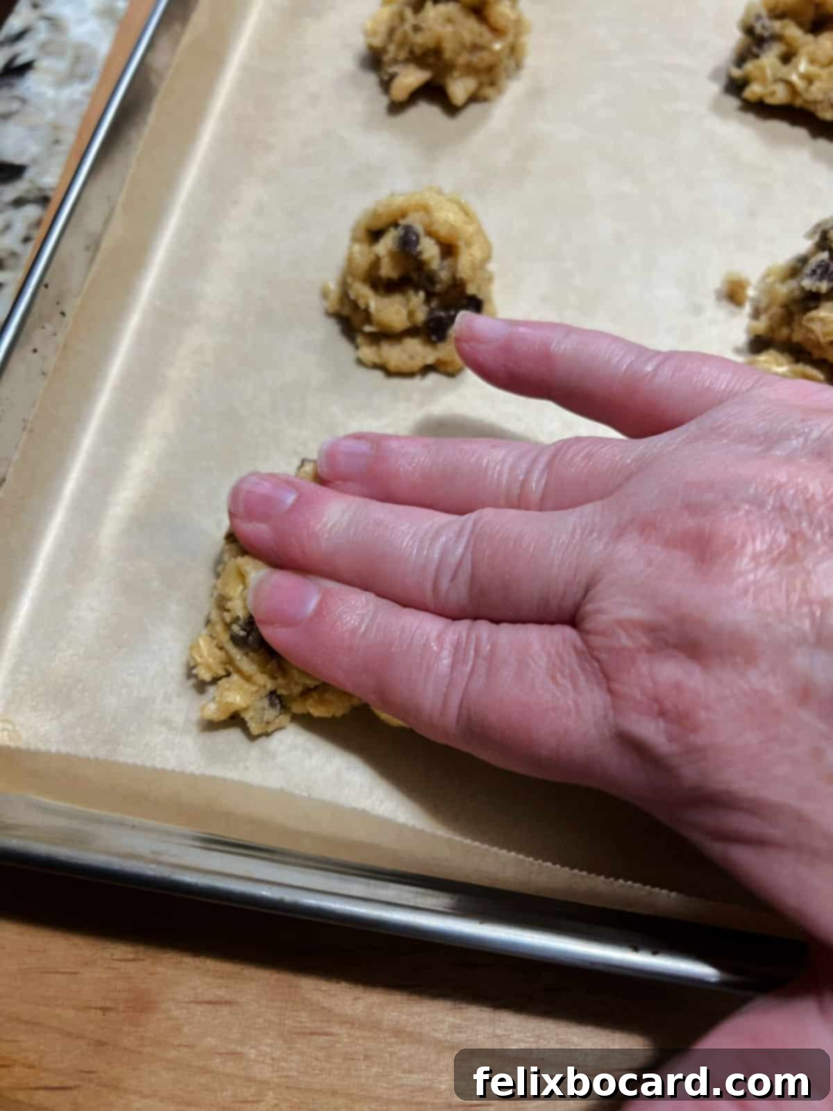 Chocolate Chip Cookies with a Crispy Secret 4 Close-up of a hand gently flattening a ball of oatmeal rice krispie cookie dough on a parchment-lined baking sheet before baking.
