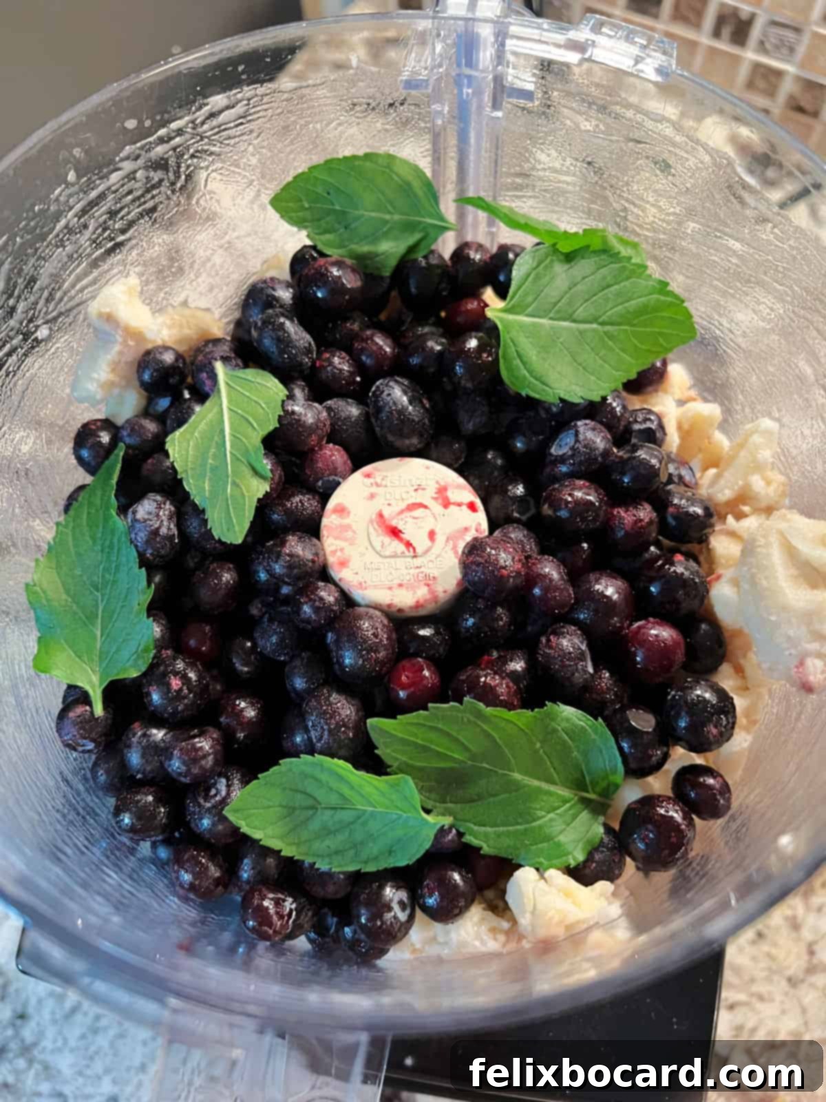 A food processor filled with frozen banana chunks, blueberries, and fresh mint leaves, ready to be blended into nice cream.