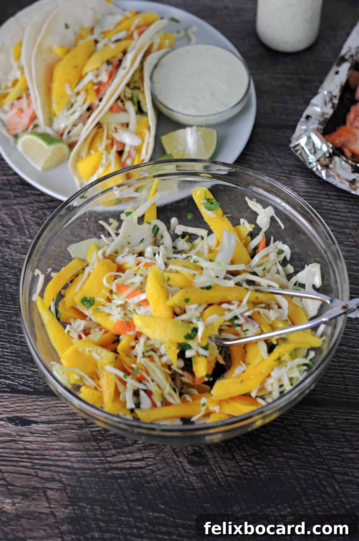 Tropical Mango Slaw for Fish Tacos 2 A vibrant bowl of fresh mango slaw sits proudly in the foreground, with a plate of tantalizing fish tacos, each adorned with the same colorful slaw, blurred elegantly in the background. The image showcases the irresistible appeal of the slaw as a perfect taco topping.