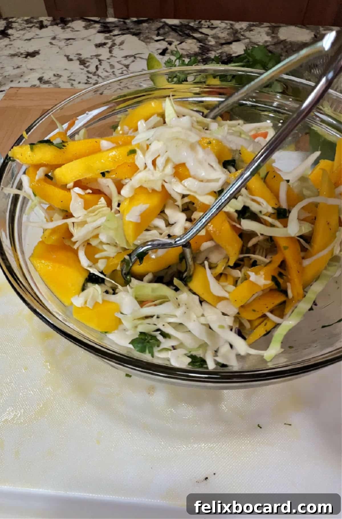 Tropical Mango Slaw for Fish Tacos 5 Tongs gently tossing to mix the vibrant mango slaw ingredients in a clear glass bowl, showcasing the fresh colors of cabbage, mango, and cilantro.