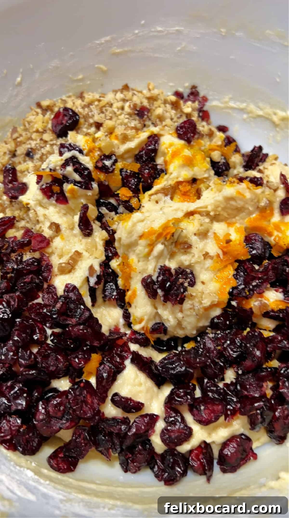 Adding dried cranberries, walnuts, and orange zest to muffin batter in a mixing bowl.