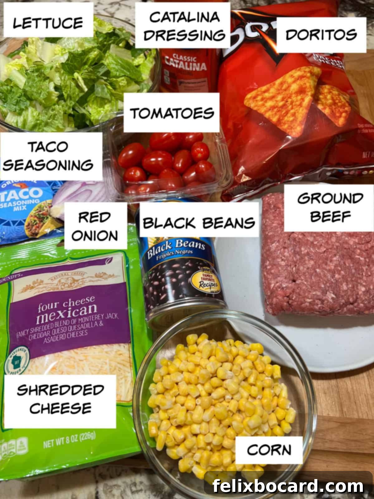 A flat lay of all fresh ingredients for Dorito taco salad: romaine lettuce, Catalina dressing, Nacho Cheese Doritos, grape tomatoes, uncooked ground beef, taco seasoning packet, sliced red onion, canned black beans, frozen corn, and shredded cheese.