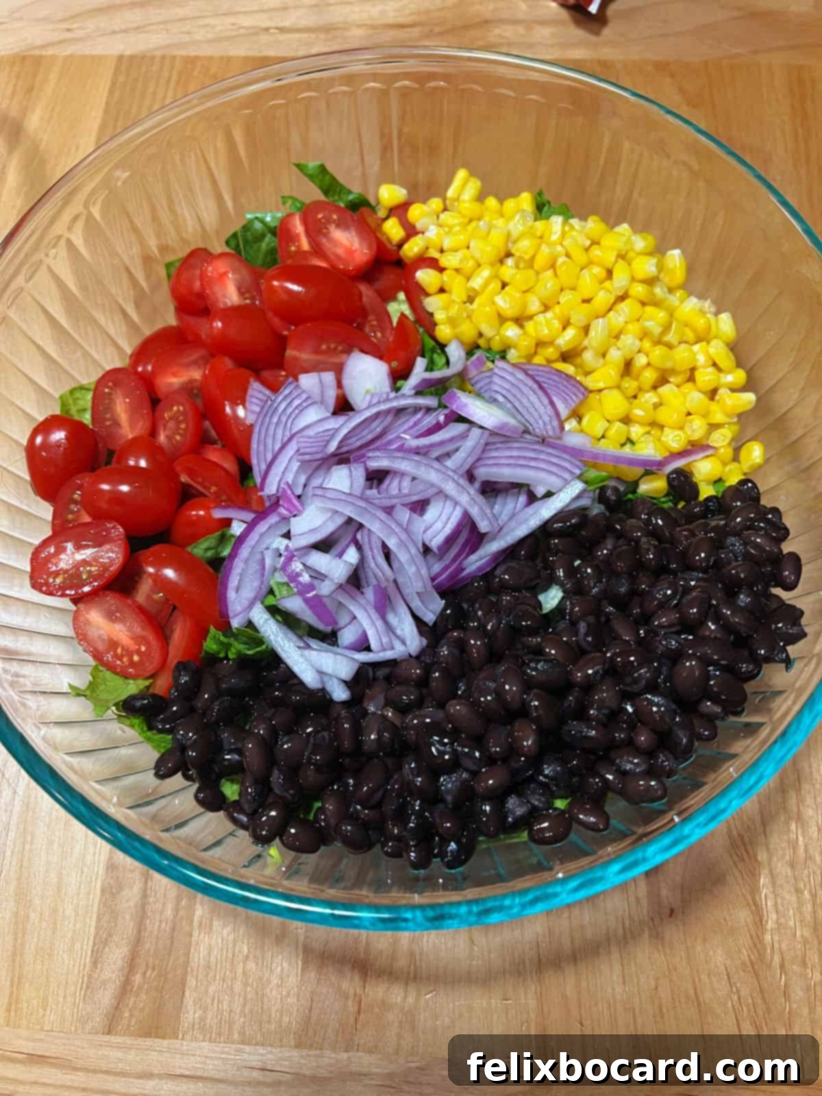 Freshly chopped lettuce, halved grape tomatoes, thawed corn, rinsed black beans, and thinly sliced red onion in a large salad bowl, ready for assembly.