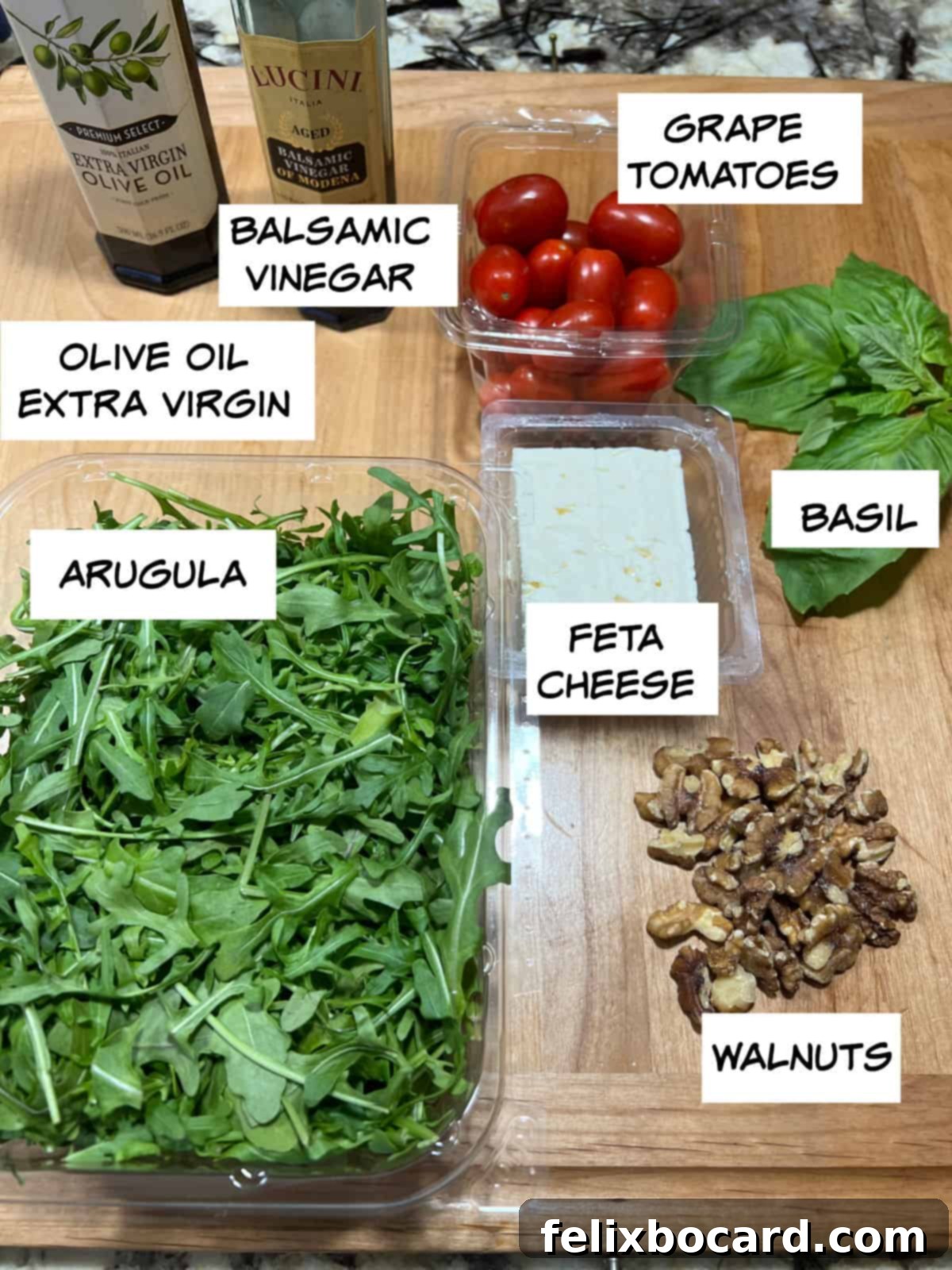 A flat lay photograph displaying all the fresh ingredients for the Arugula Feta Tomato Salad: arugula, olive oil, balsamic vinegar, tomatoes, basil, feta cheese, and walnuts.