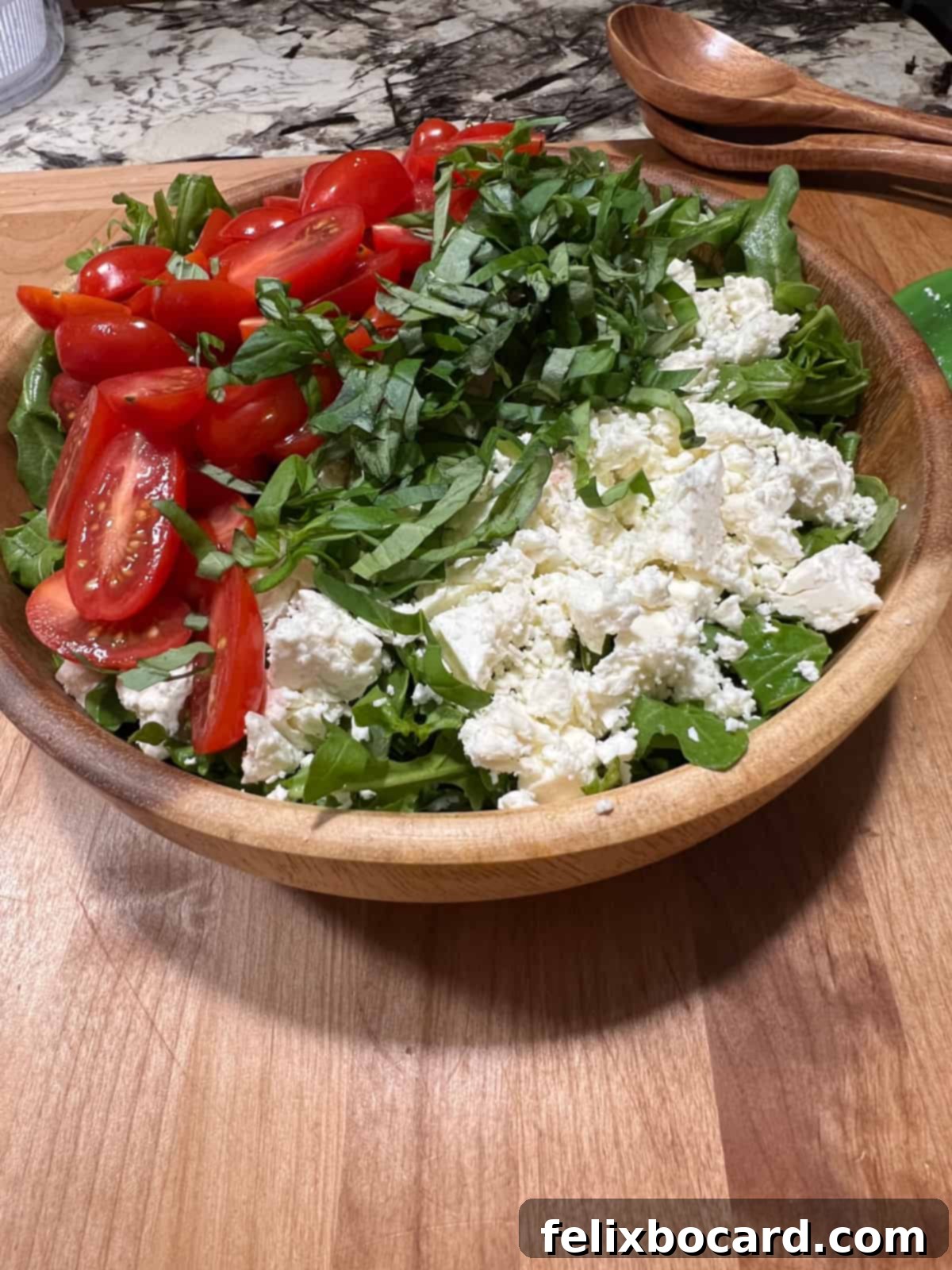 A wooden bowl filled with undressed salad ingredients: arugula, tomatoes, and feta cheese.