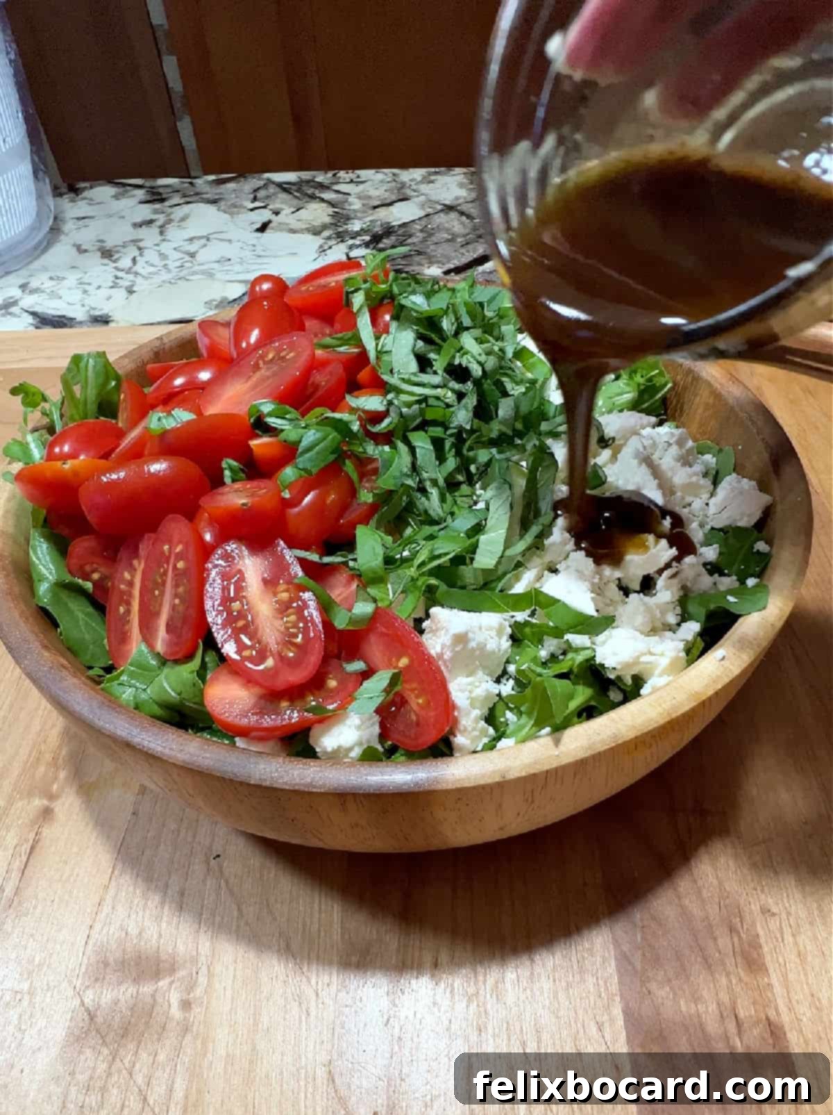 A small cup of balsamic dressing being poured over the mixed salad ingredients in a bowl.