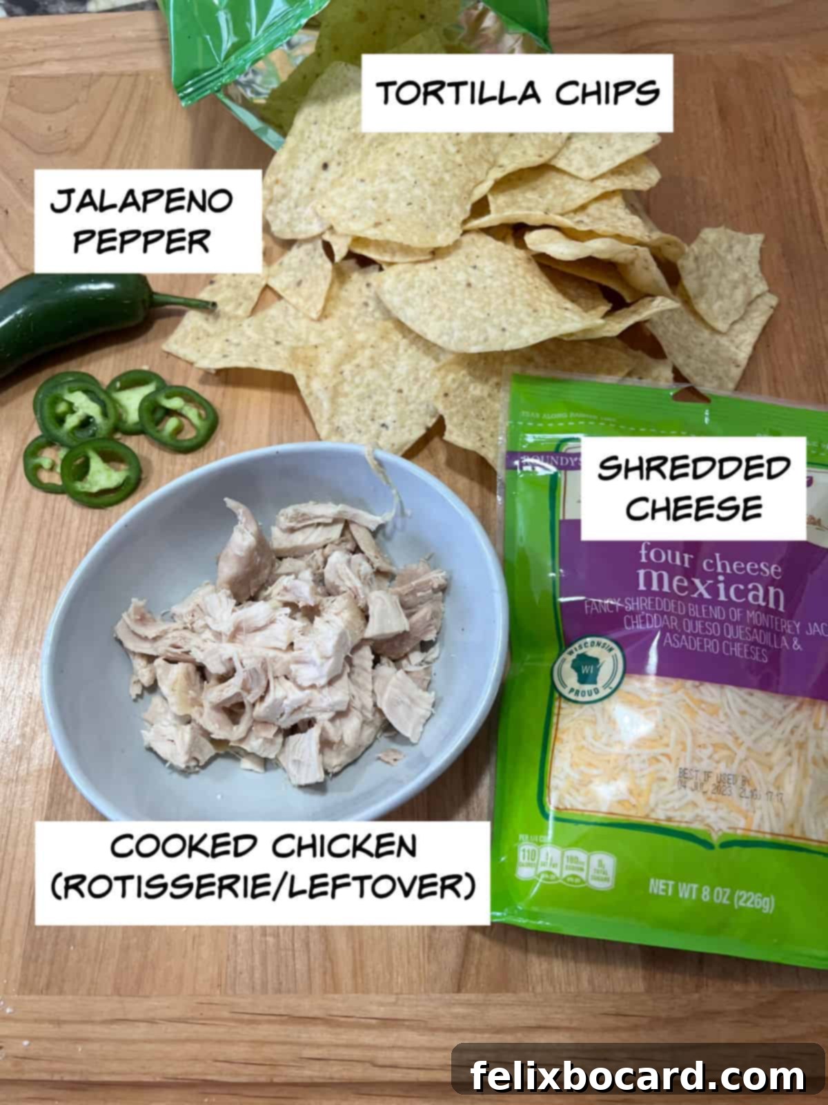 Ingredients for air fryer nachos arranged on a cutting board: a bowl of tortilla chips, shredded cheese, cooked shredded chicken, and sliced jalapeno pepper.