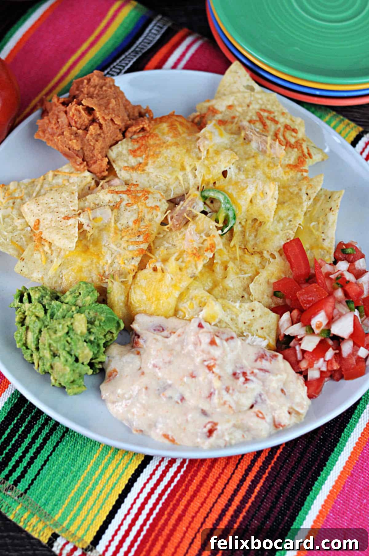 A vibrant plate of loaded nachos surrounded by bowls of refried beans, guacamole, creamy queso dip, and fresh salsa for dipping.