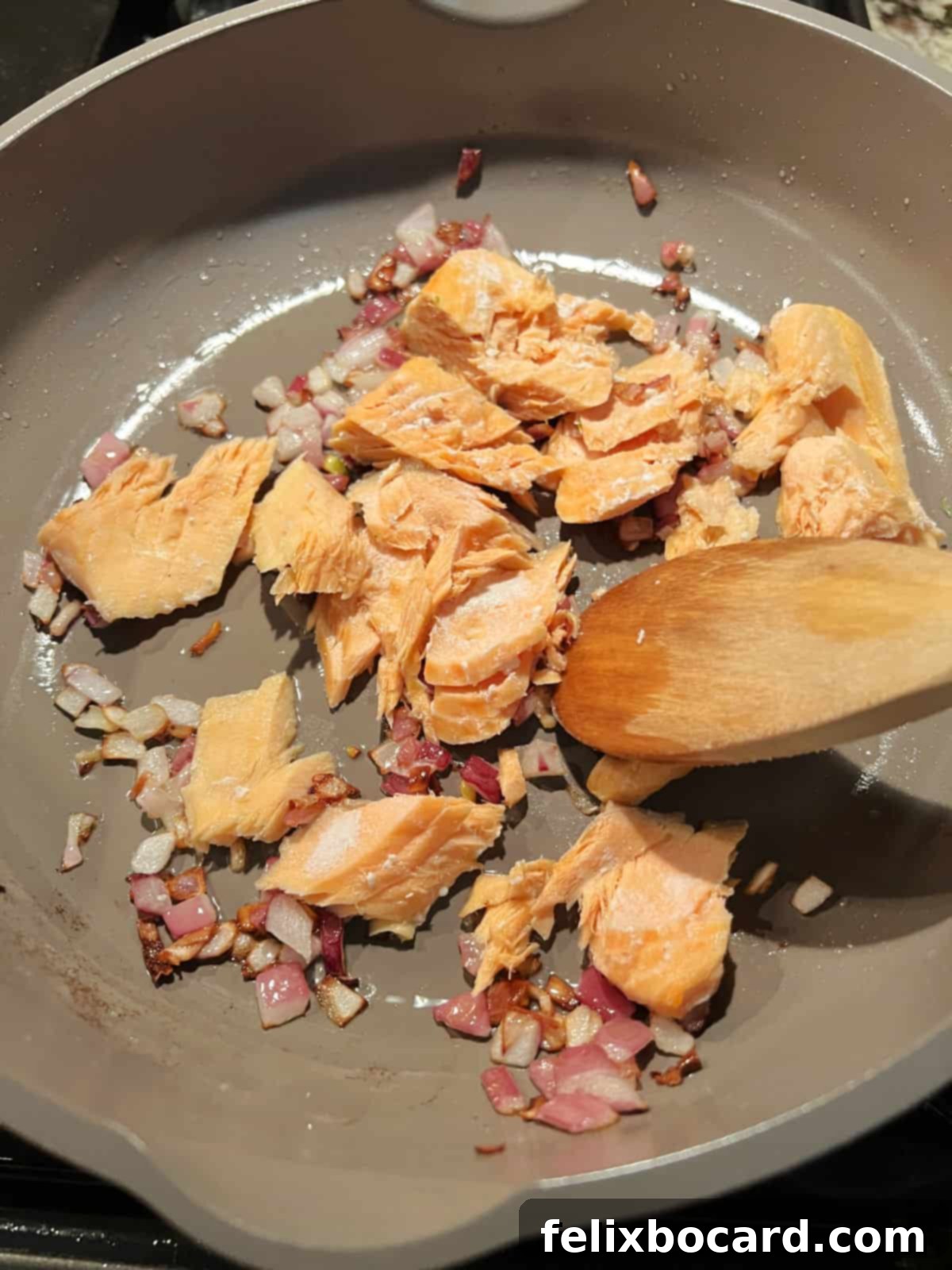 A close-up shot of cooked salmon being flaked apart in a skillet with a wooden spoon, alongside sautéed red onions.
