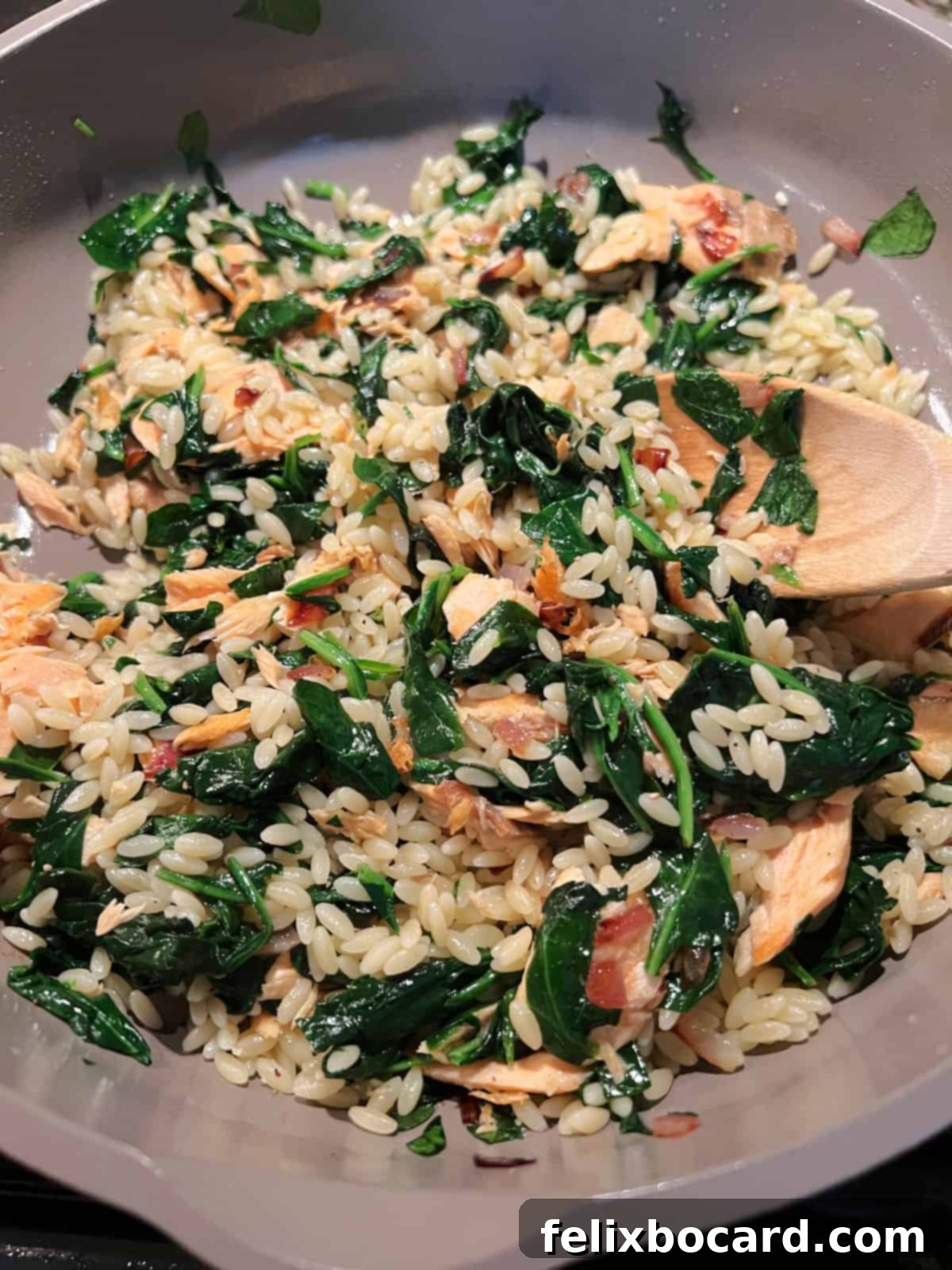 A skillet filled with wilted spinach and cooked orzo pasta, mixed with flaked salmon and sautéed onions.
