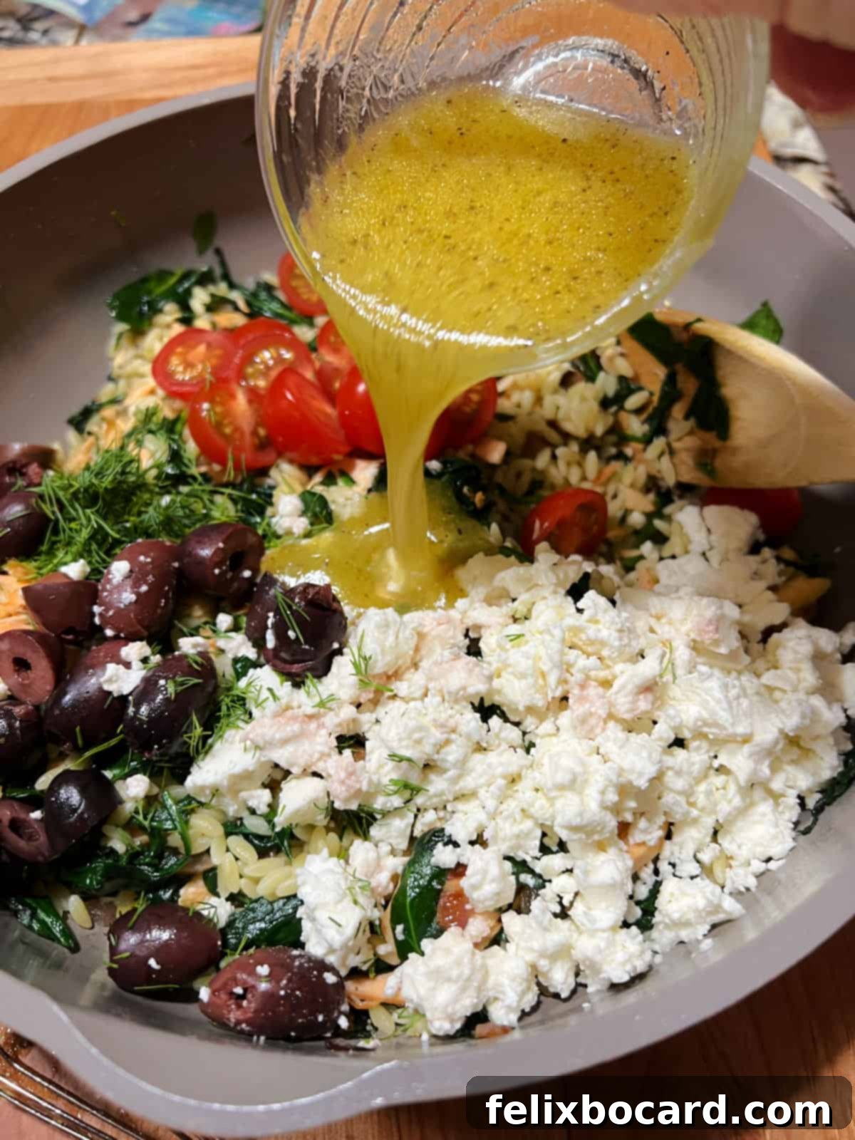 A hand pouring a lemon-dill dressing over a colorful Salmon Orzo Salad in a large skillet, ready for final mixing.