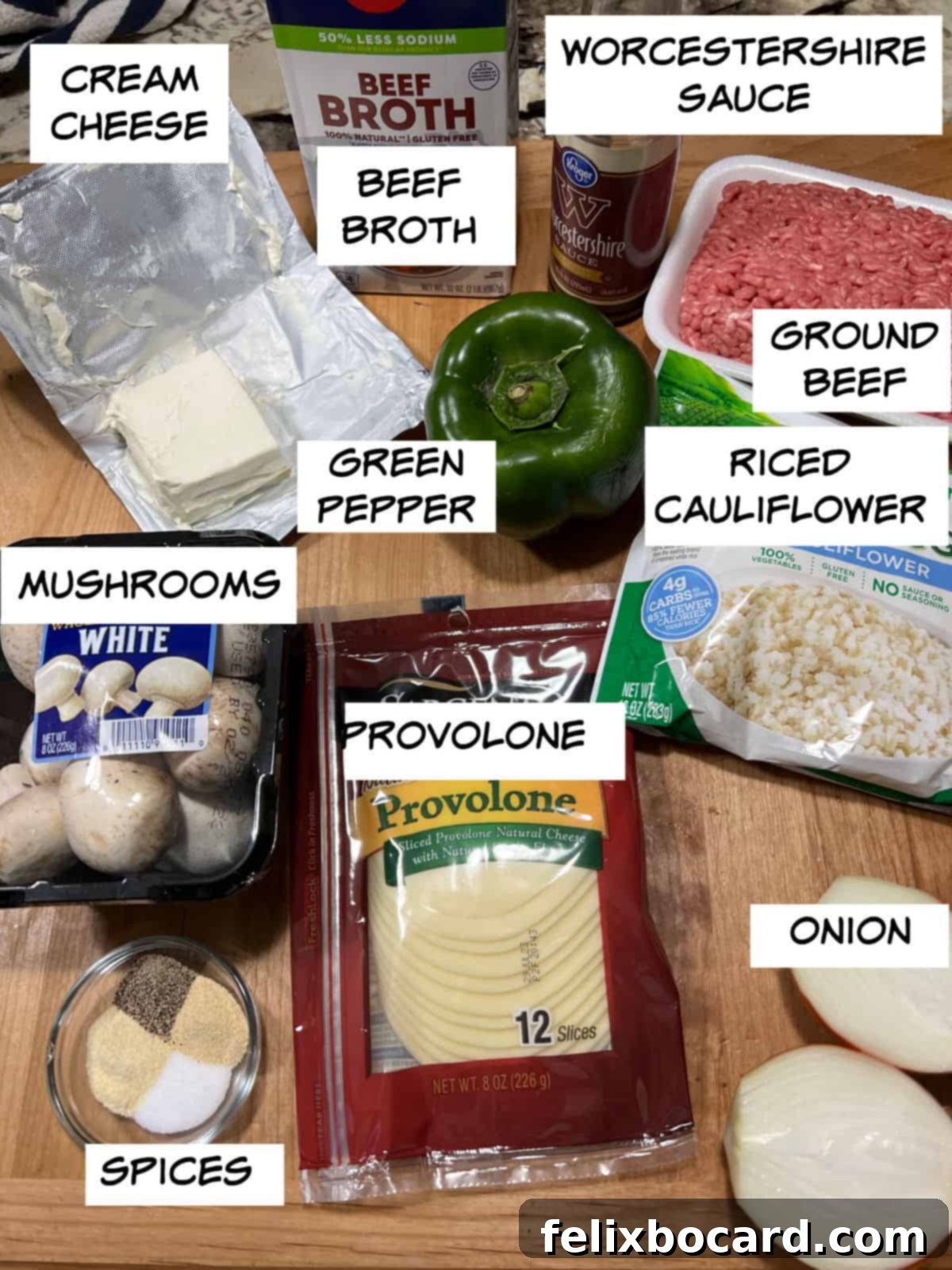 A flat lay of fresh ingredients for Philly Cheesesteak Casserole, including cream cheese, beef broth, Worcestershire sauce, ground beef, riced cauliflower, onion, green pepper, mushrooms, provolone cheese, and various spices.