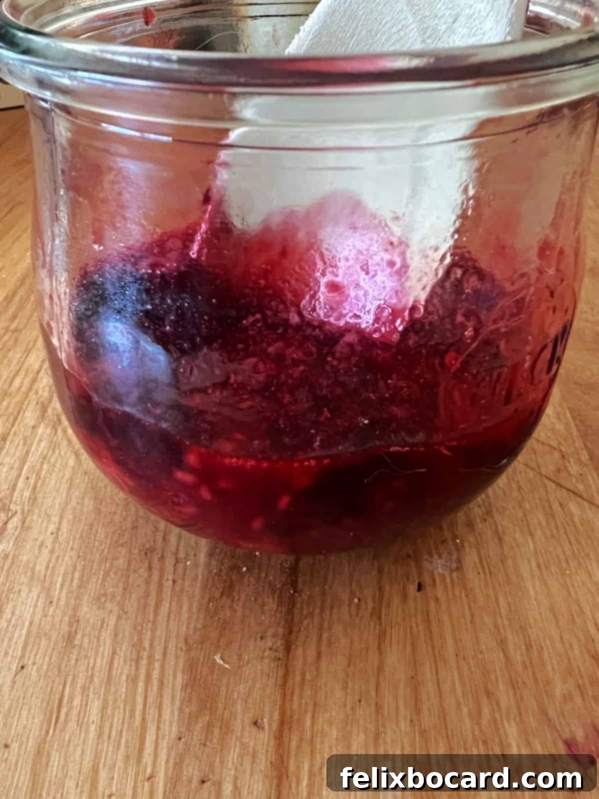 A wooden spoon mashing thawed berries against the bottom of a glass jar, releasing juices.