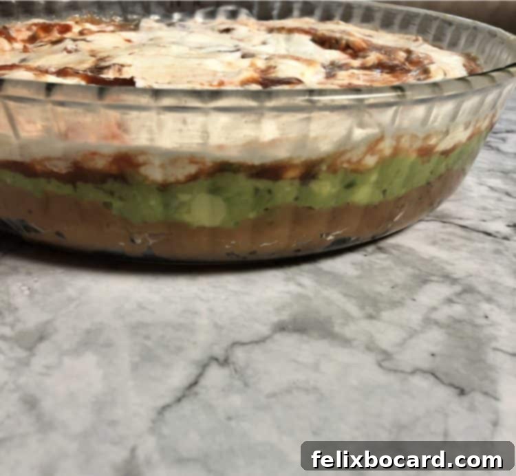 A beautifully layered taco dip presented in a clear, round glass pan, showcasing distinct layers of beans, guacamole, salsa, and sour cream.