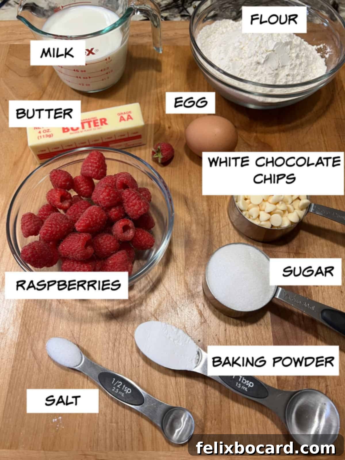 A flat lay of all ingredients laid out: a bowl of milk, melted butter in a ramekin, a single egg, a pile of flour, a small bowl of sugar, baking powder, salt, a bowl of white chocolate chips, and a pint of fresh raspberries.