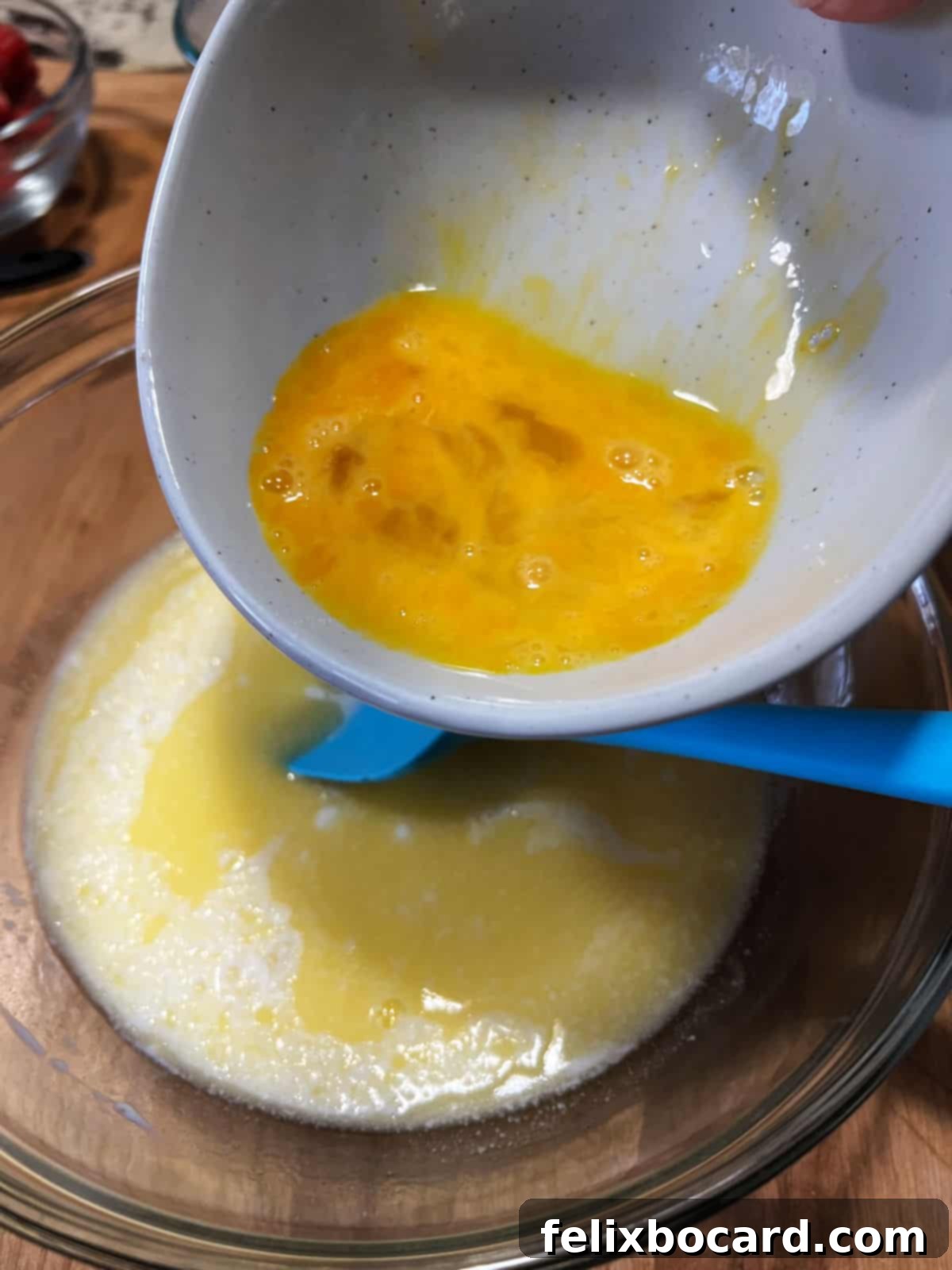 A close-up shot of a whisk combining a slightly beaten egg with melted butter and milk in a glass bowl.