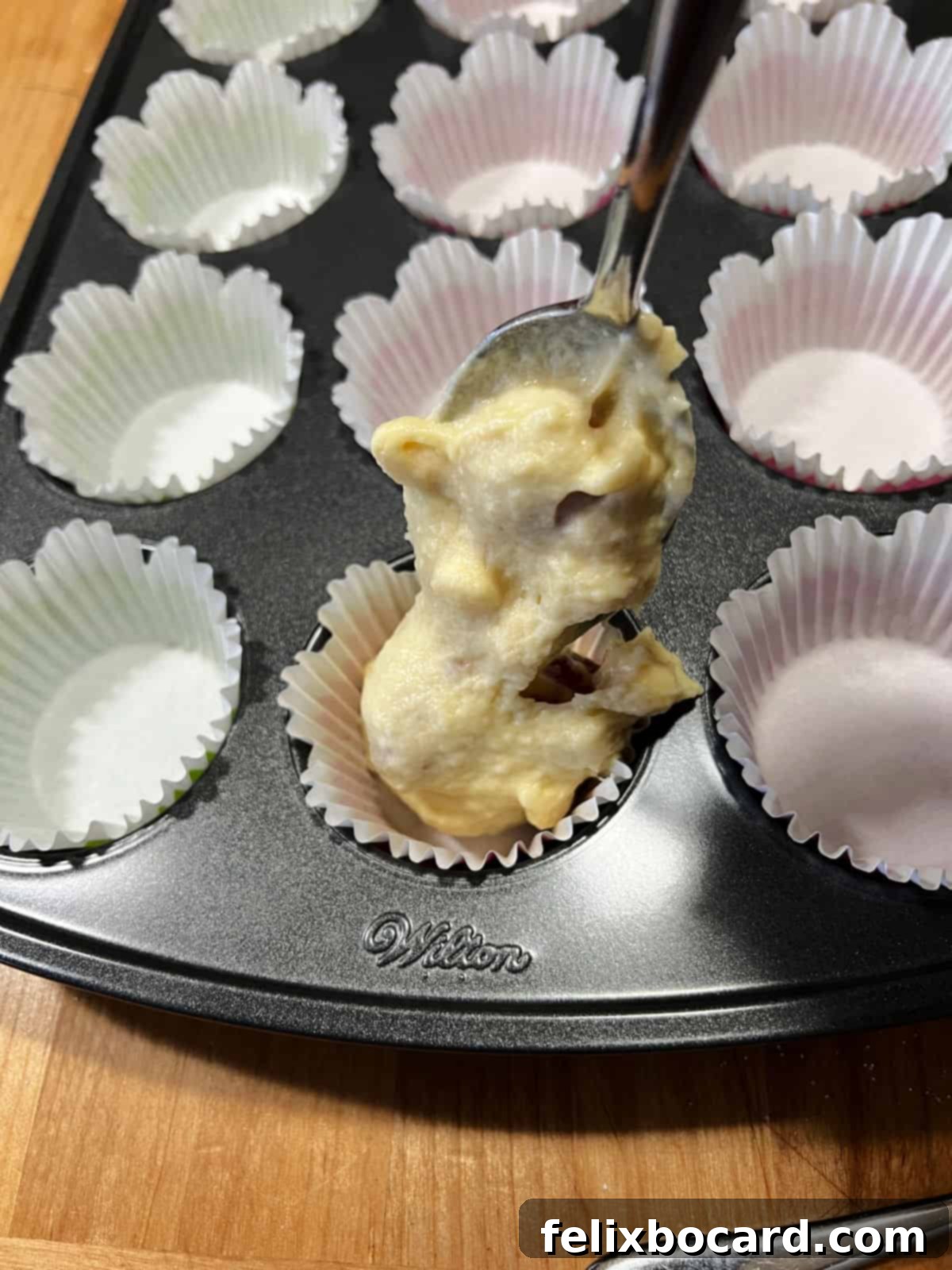 A spoon full of muffin batter being carefully placed into a paper-lined muffin cup in a baking pan.